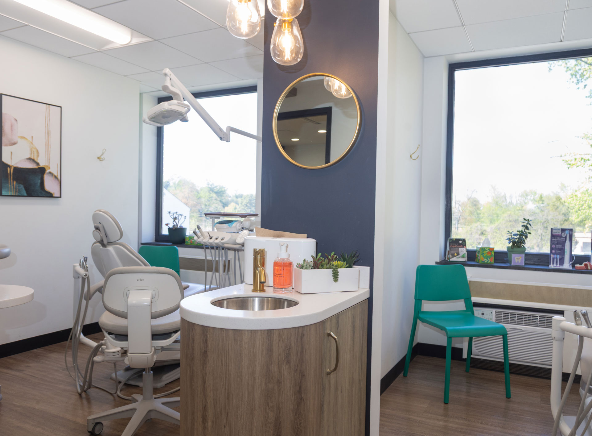 side by side exam rooms with dental chairs and equipment facing windows looking outside and cabinet, sink, and mirror on wall in between